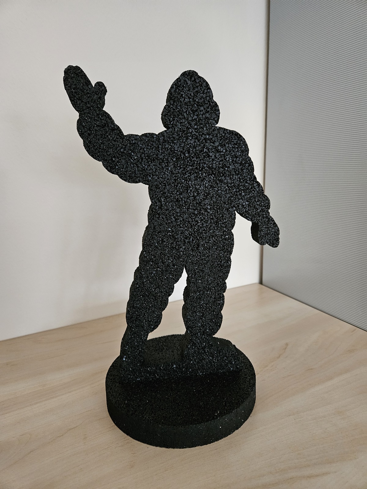 Schwarze Kunstfigur aus Granulat auf rundem Sockel, abstrakte menschliche Form mit erhobener Hand, moderne Skulptur.