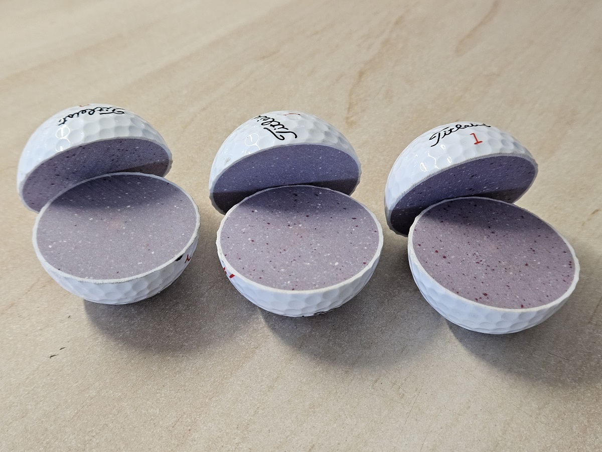 Drei geöffnete Titleist Golfbälle mit grauem Pulver gefüllt, auf beiger Oberfläche angeordnet.