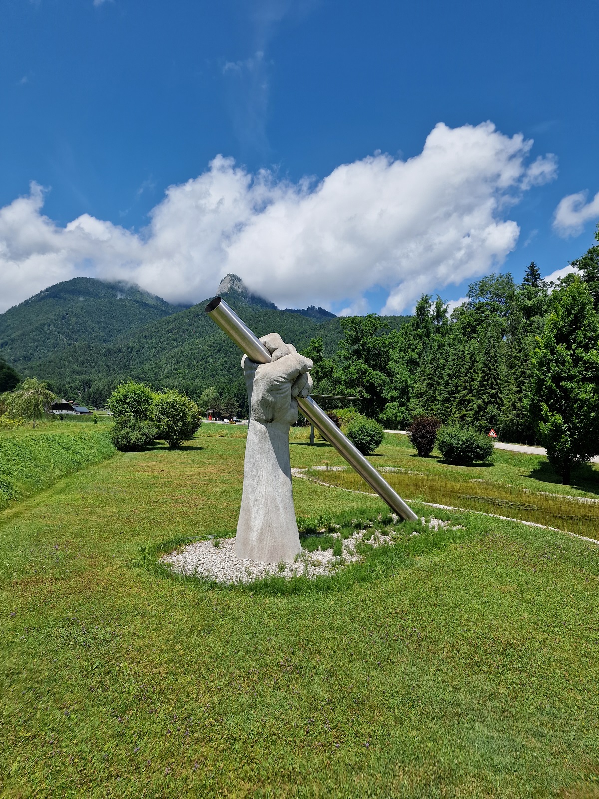 Edelstahl-Kunstobjekt: Figur hält großes Stahlrohr in alpiner Berglandschaft mit grüner Wiese und Waldkulisse.