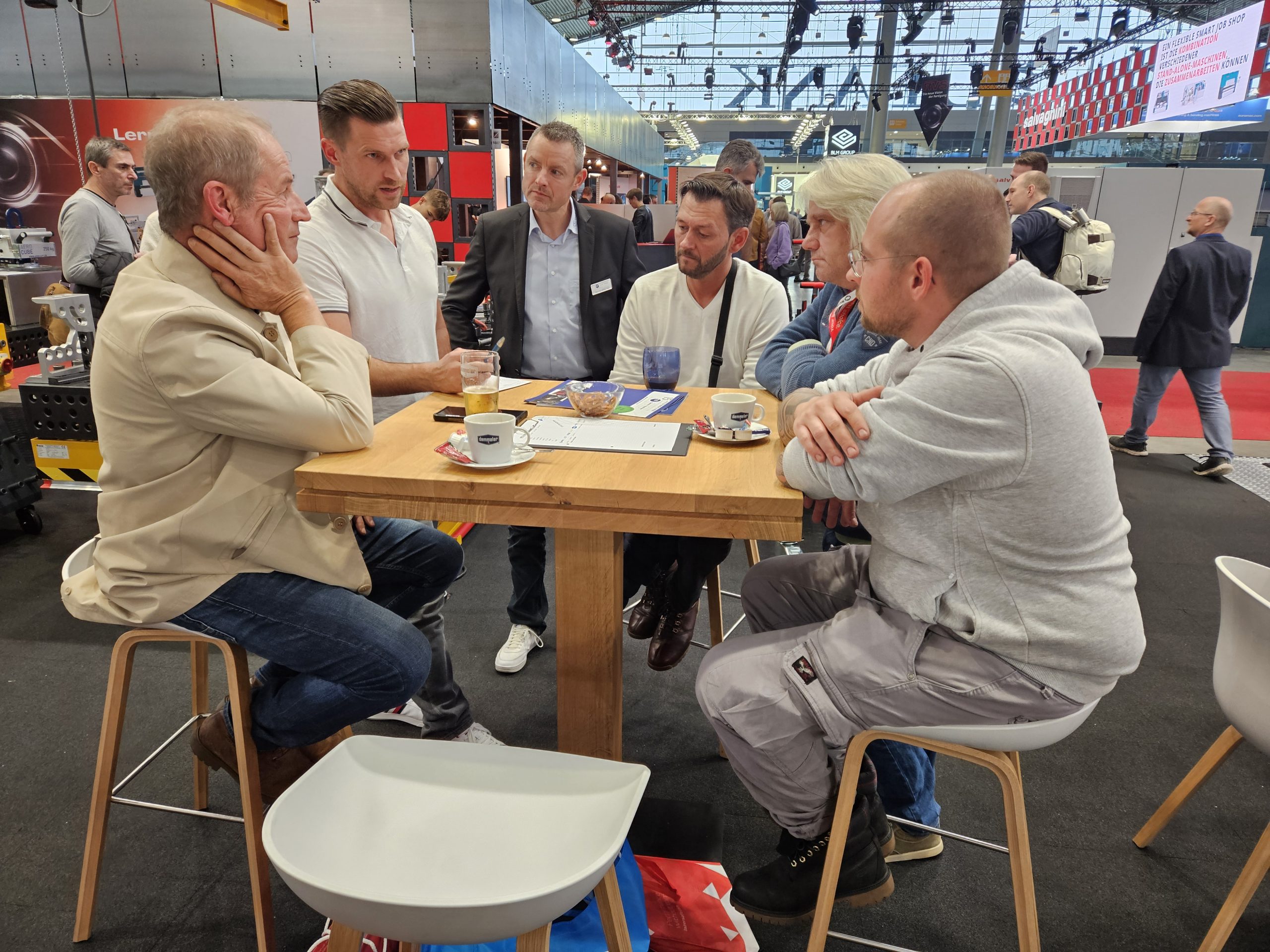 Geschäftsmänner und Besucher beim Austausch an Holztisch auf Messe mit Kaffee und Getränken, Hallenambiente im Hintergrund.