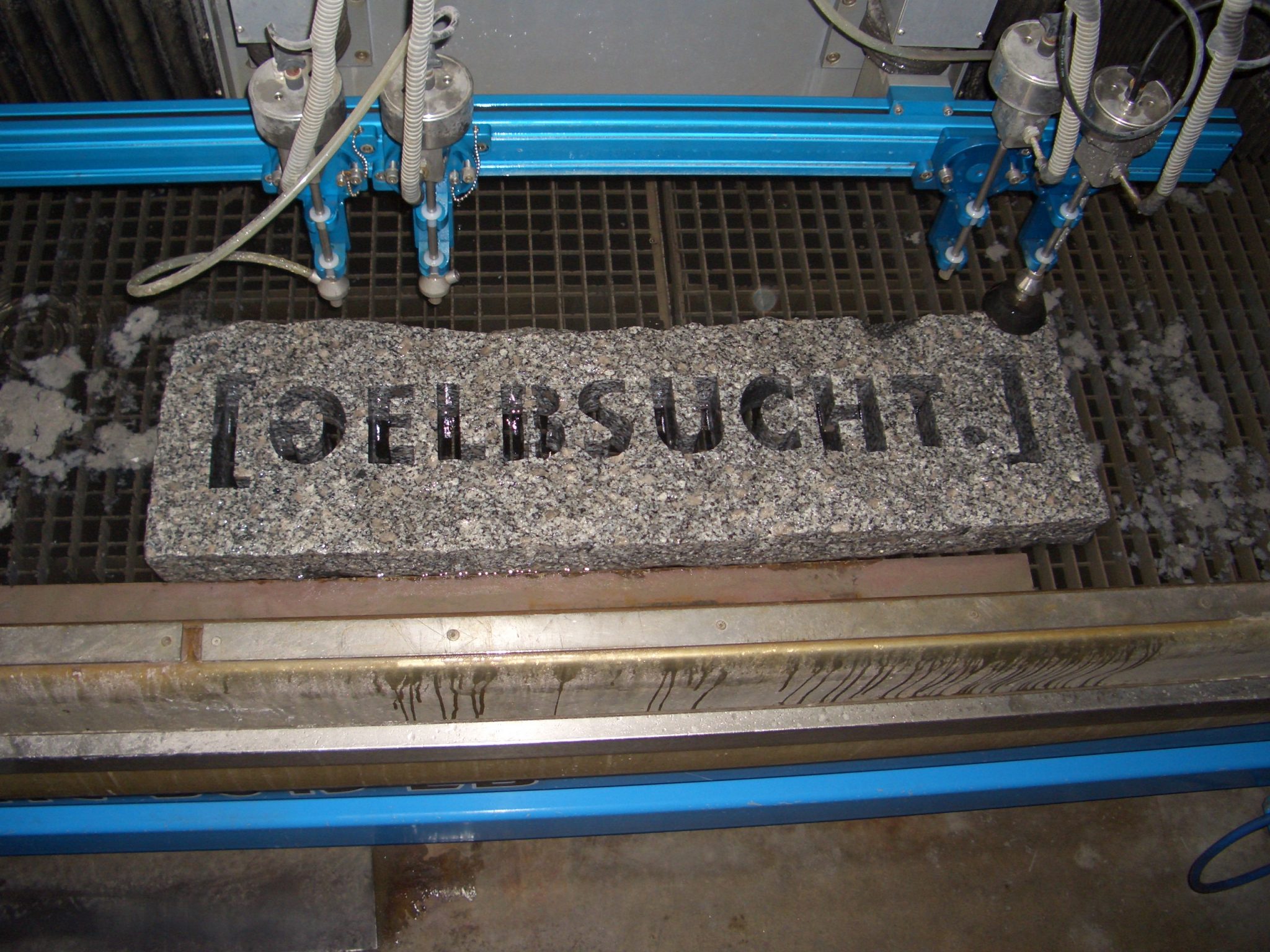 Wasserstrahlschnitt auf Granit-Naturstein mit eingravierten Buchstaben, montiert auf blauem Schneidtisch mit Düsensystem.