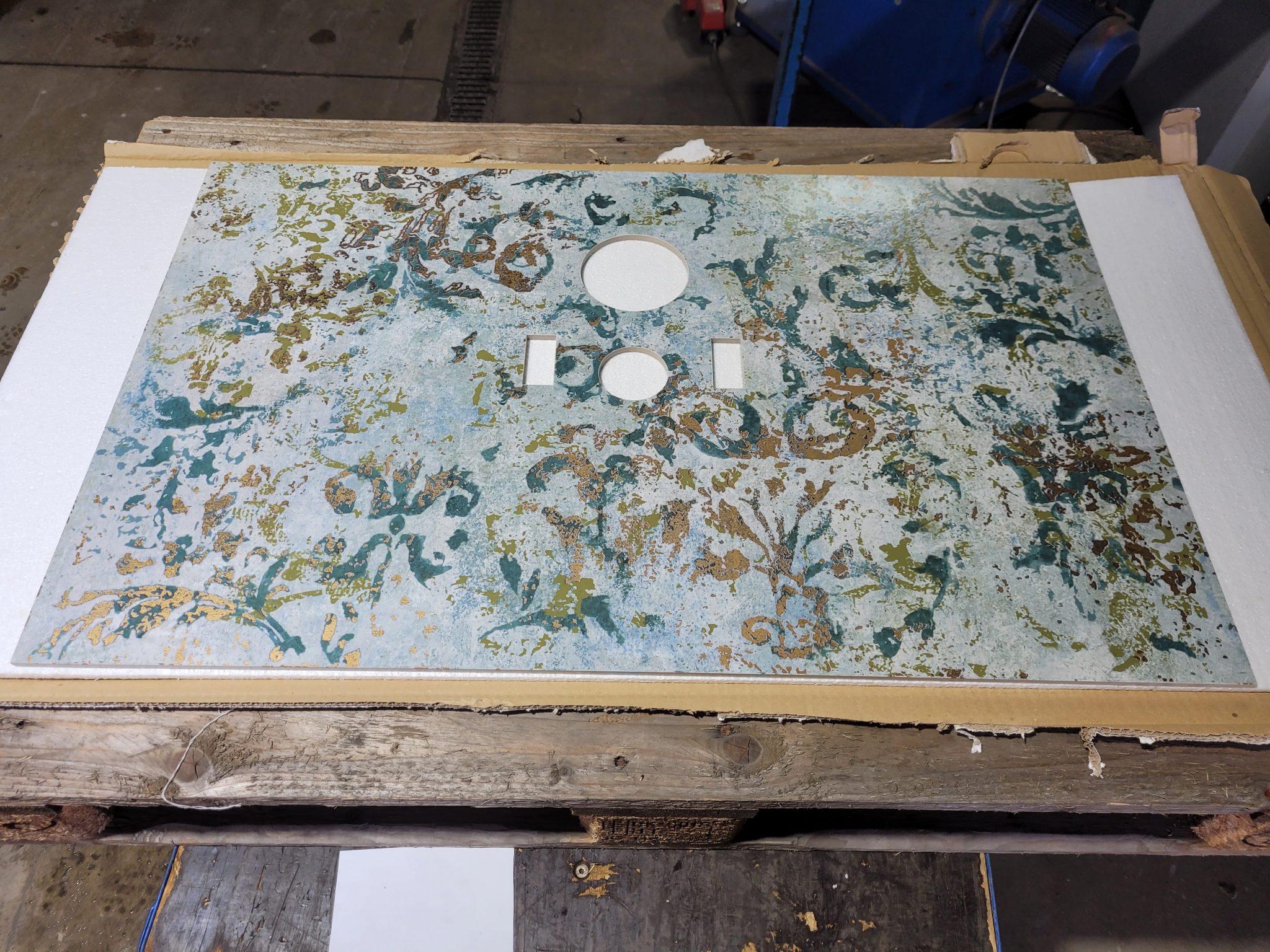 Edelstahlplatte mit abstraktem Oberflächendesign in Grün- und Goldtönen, Bohrungen für Montage, auf Holzpalette.