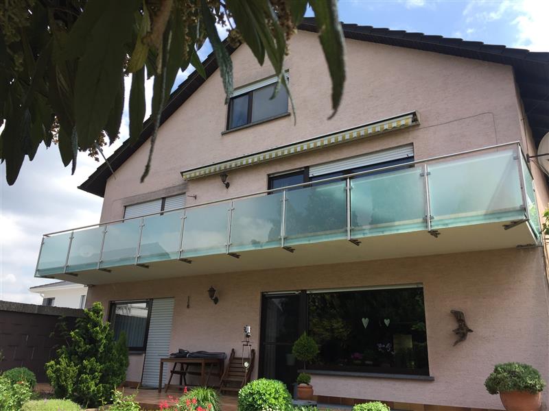 Modernes Wohnhaus mit Edelstahl-Glasgeländer auf großzügiger Terrasse, umgeben von Grünflächen und Pflanzen.