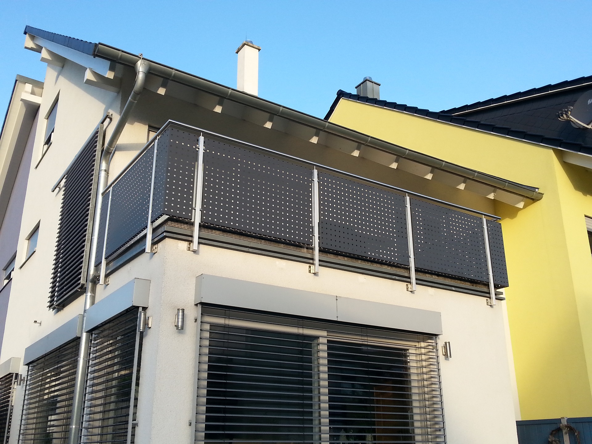 Modernes Wohnhaus mit Edelstahl-Balkongeländer und perforiertem Sichtschutz, zeitgenössische Architektur mit Jalousien und gelber Fassade.