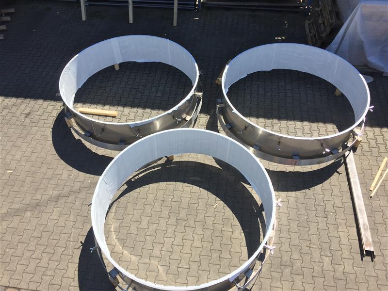 Drei metallische Edelstahl-Rohre mit weißen Kunststoffringen auf gepflastertem Untergrund, Werkstatt-Setting für Edelstahlverarbeitung.