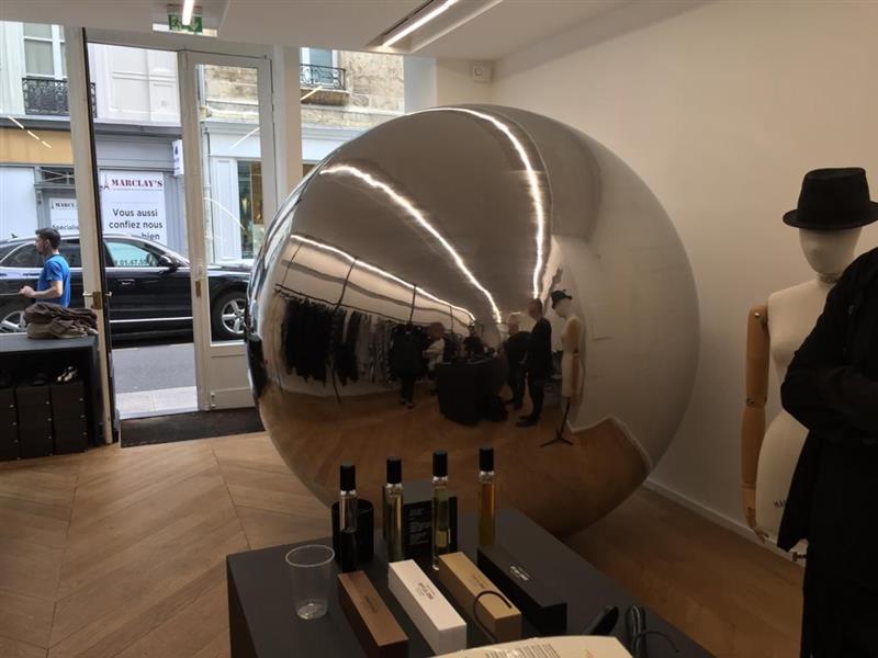 Glänzende Edelstahlkugel in modernem Showroom mit Spiegelung von Besuchern und Produktdisplay darunter, Schaufenster im Hintergrund.