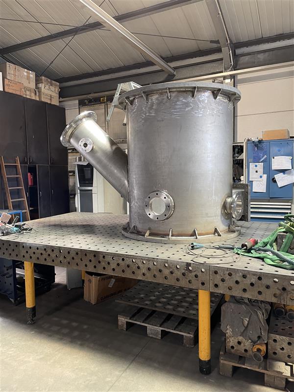 Großer Edelstahl-Druckbehälter mit Anschlussrohr auf Werkstatt-Arbeitstisch in Industriehalle mit Werkzeugen.