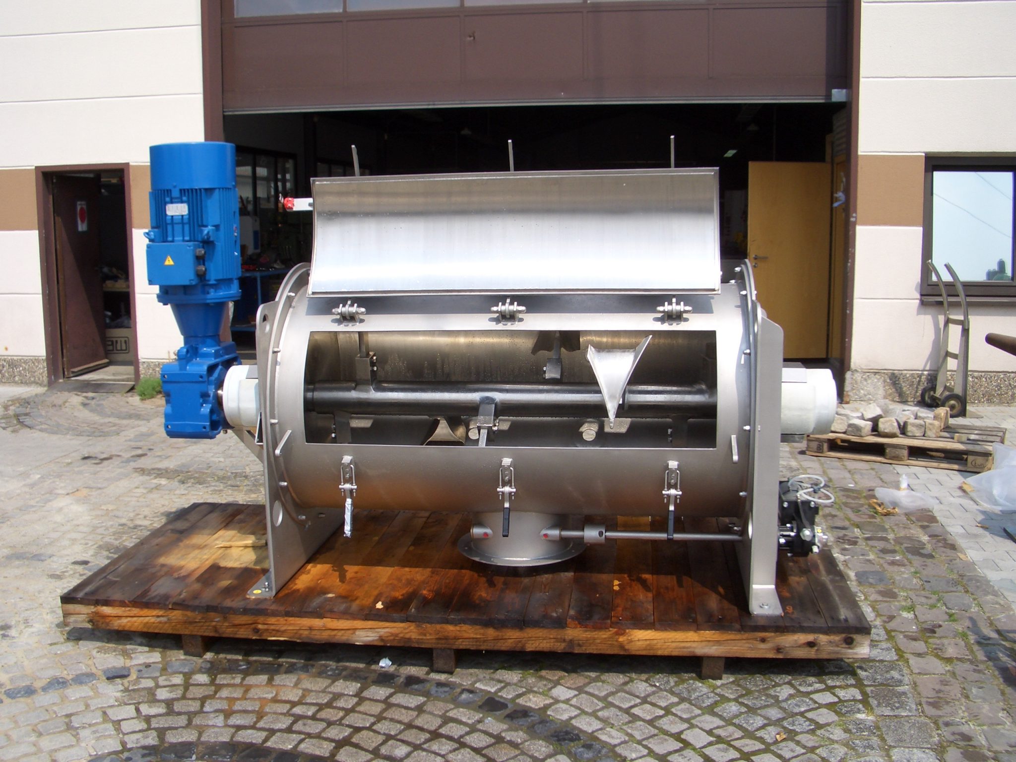 Industrielle Edelstahl-Mischtrommel mit blauem Elektromotor auf Holzpalette in Fabrikhof montiert.