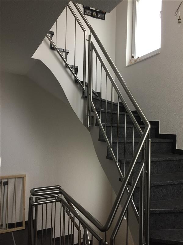 Moderne Edelstahl-Treppe mit horizontalen Stahlseilen als Geländer in hellem Treppenhaus mit schwarzen Stufen und Fenster.