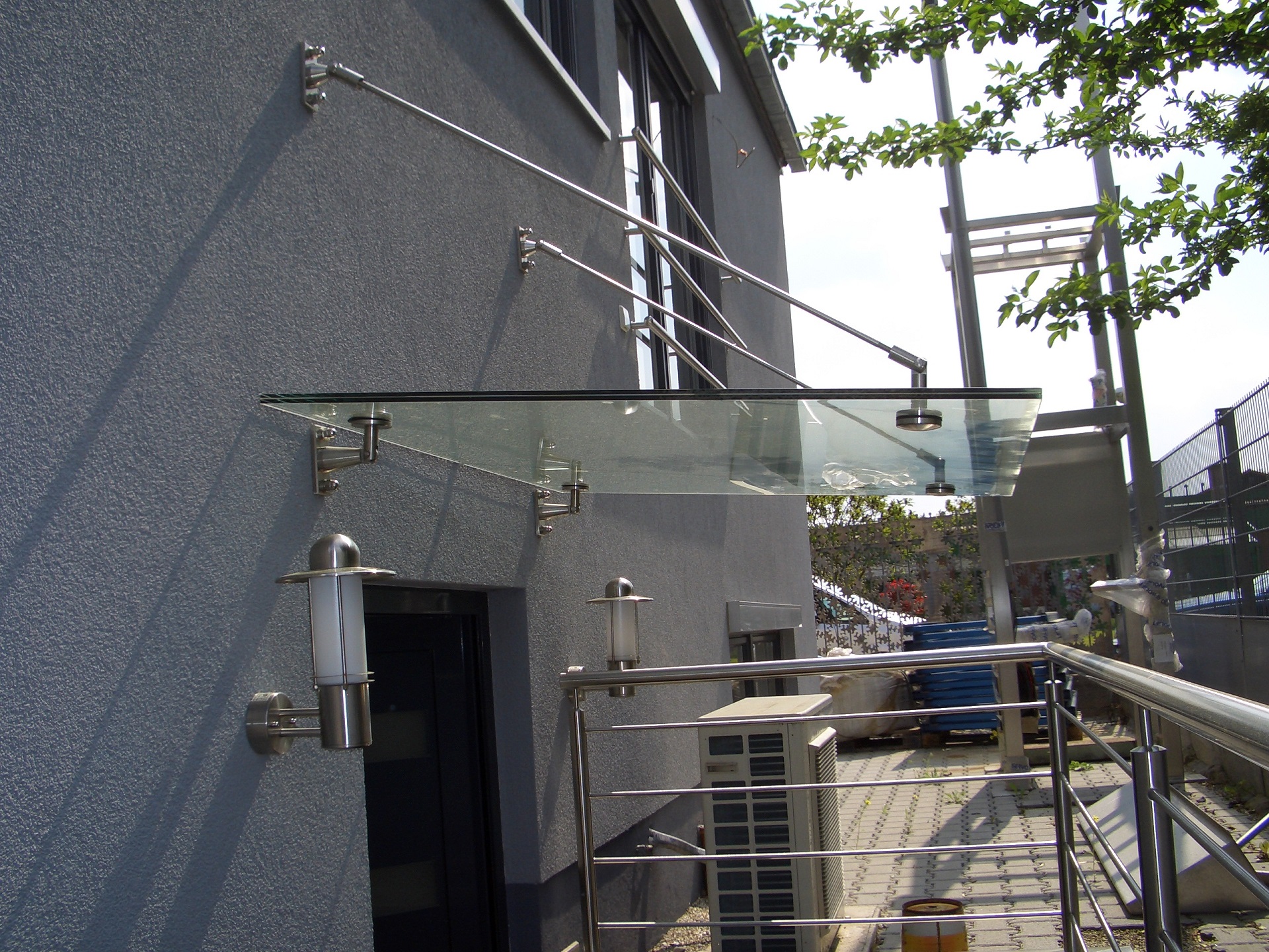 Moderne Edelstahl-Terrassenüberdachung mit Glaselementen, Geländer und Wandleuchten an grauer Fassade.