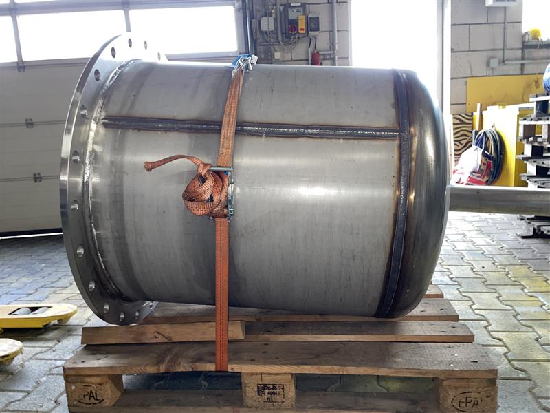 Großer Edelstahl-Druckbehälter auf Holzpalette, gesichert mit orangefarbener Spanngurt, in Industriewerkstatt.