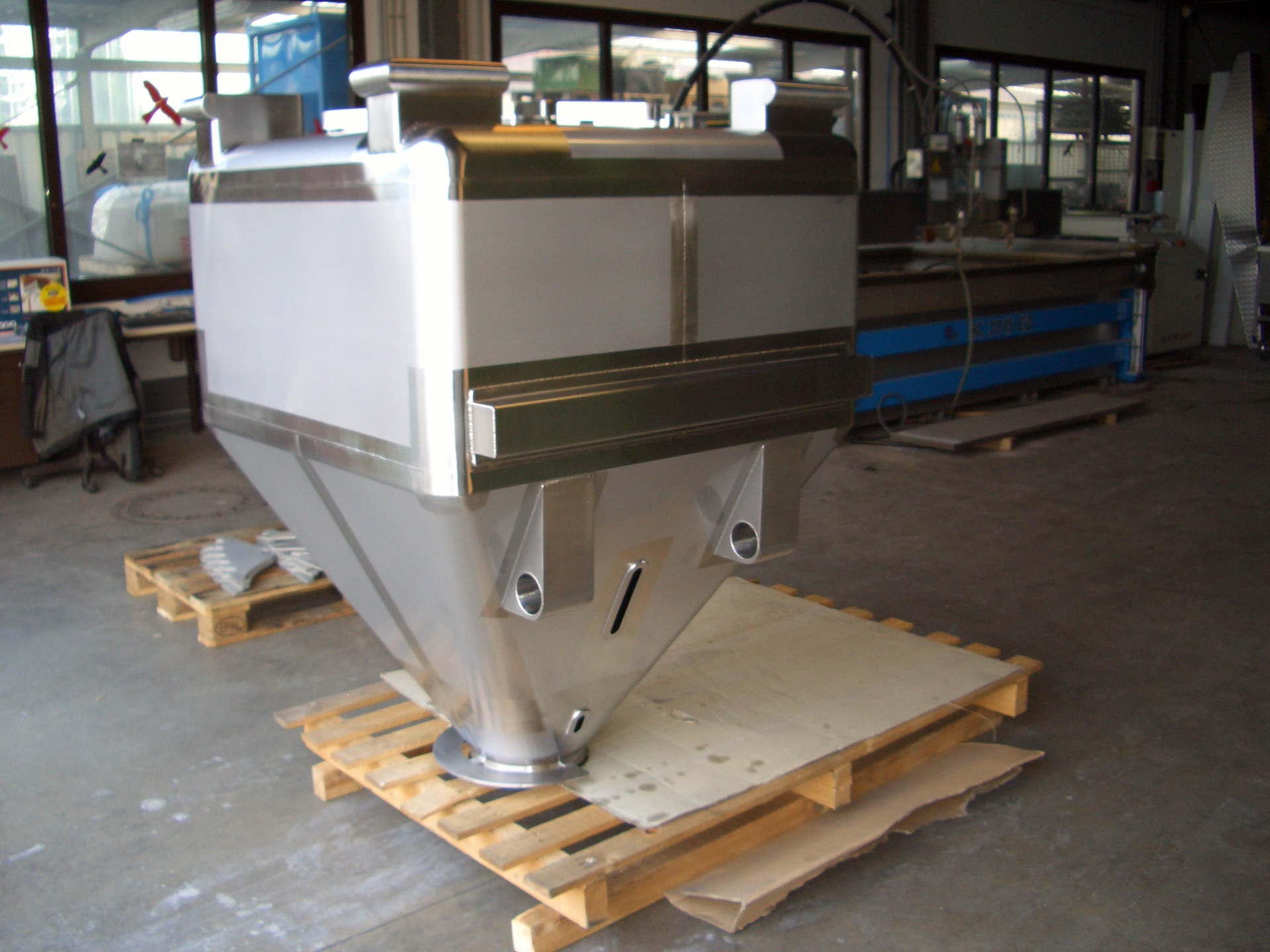 Edelstahl-Wassertank mit braunem Deckel auf Holzpaletten in Produktionshalle, Wasserstrahlschneidanlage im Hintergrund.