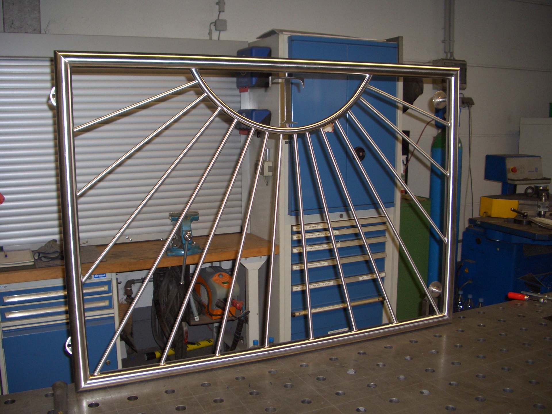 Edelstahl-Schiebetür mit Sonnendesign in Werkstatt, dahinter Arbeitsgeräte und blaue Werkstatteinrichtung von Klumpp.
