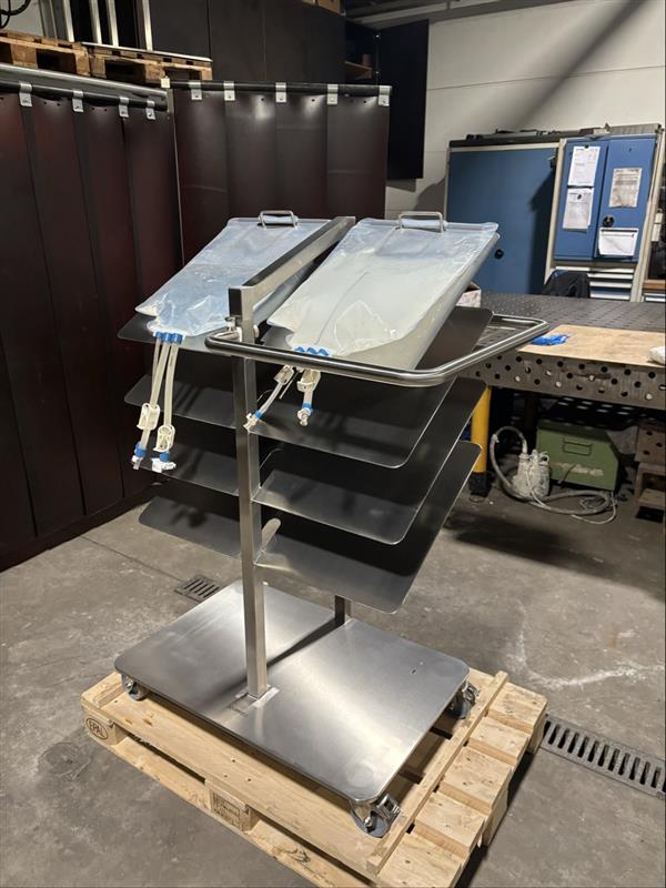 Edelstahl-Arbeitstisch mit gekippbaren Tabletts auf Rädern, Werkstatt mit Schutzvorhang und Werkzeugen.
