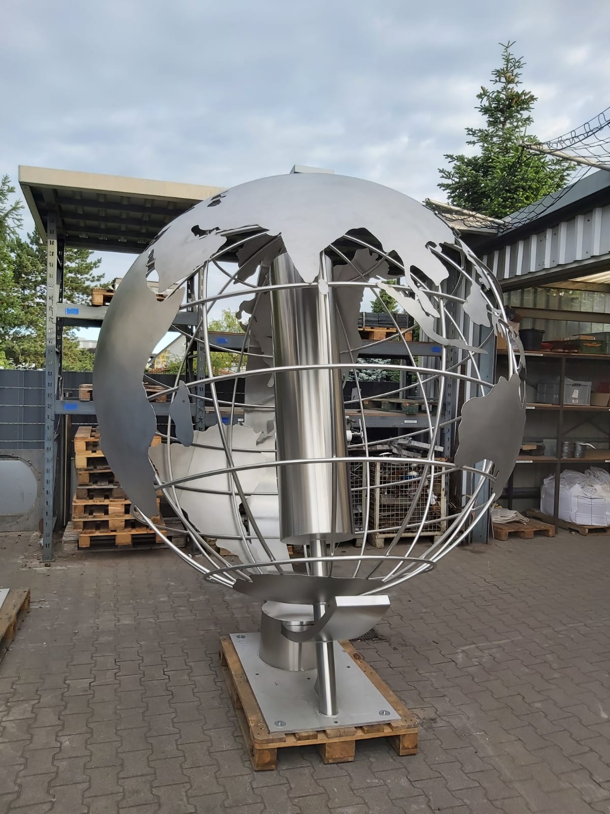 Große Edelstahlkugel mit geometrischem Gittermuster und zentralem Rohr, montiert auf Sockel in Industriehof.