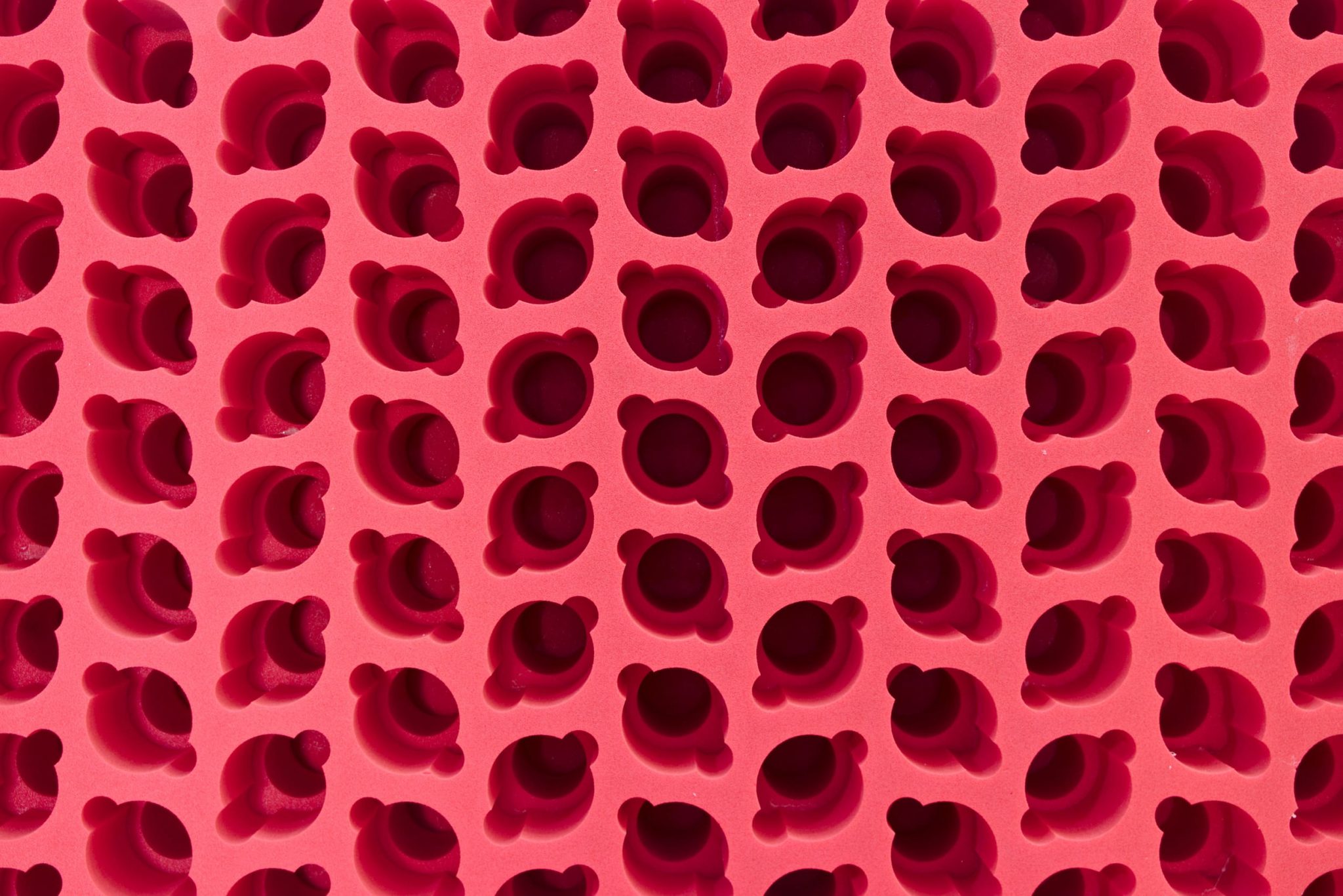 Repetitives Muster aus dunkelroten 3D-Objekten auf rosa Hintergrund mit Schattenwurf, abstrakte geometrische Oberflächenstruktur.
