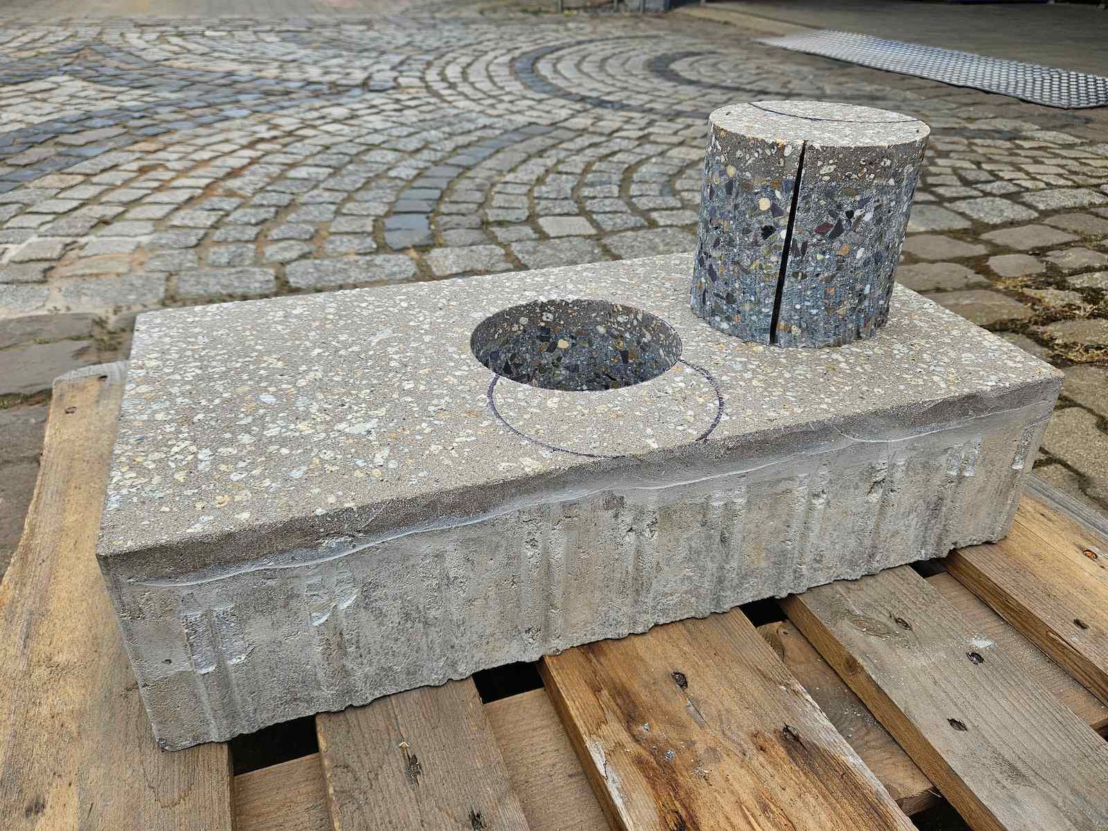 Betonwerkstück mit Durchbruch und Holzpfosten auf Holzpalette, gepflasterter Hof im Hintergrund.